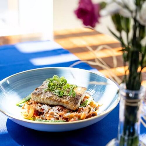 Filet z ryby na makaronie w sosie pomidorowym z mikroliśćmi, podany w niebieskiej misce na stoliku jasnej restauracji hotelowej.