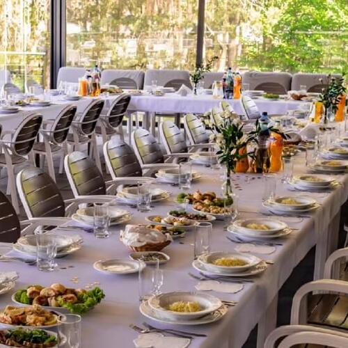 Bankietowy stół w restauracji z białymi obrusami, ustawionymi talerzami, napojami i widokiem na ogród.