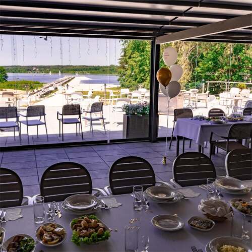 Taras restauracyjny z nakrytym stołem i złoto-białymi balonami, przeszklona weranda pod pergolą, widok na molo, plażę i jezioro z żaglówkami