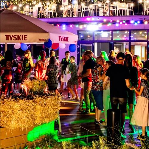 Nocna impreza na hotelowym tarasie z barem i parasolami TYSKIE, migoczącymi lampkami, stolikami i tańczącymi gośćmi.