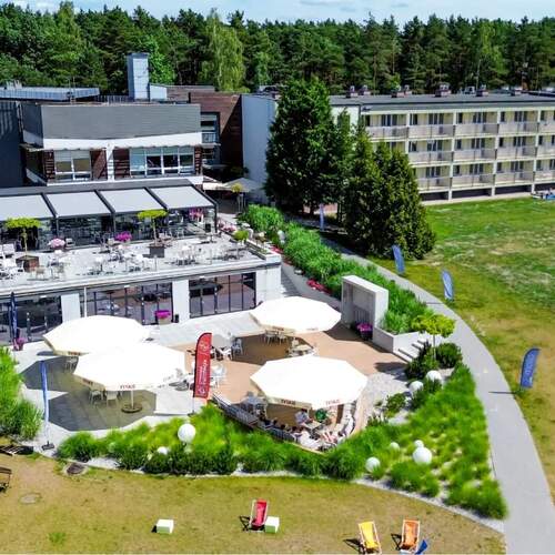 Hotel z tarasem i ogródkiem letnim, zewnętrzna restauracja pod parasolami, leżaki na trawniku, balkony pokoi i sosnowy las.