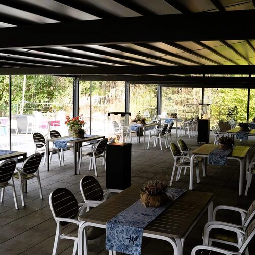 Zadaszony taras restauracyjny z stolikami, dekoracyjnymi bieżnikami, donicami, podgrzewaczami i widokiem na zielony ogród.