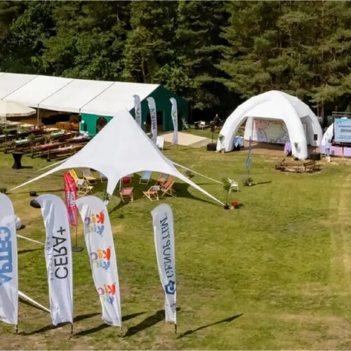 Plenerowy teren wydarzeń przy jeziorze: namioty eventowe i pawilony, strefy wypoczynku, dmuchańce dla dzieci i ławki piknikowe.