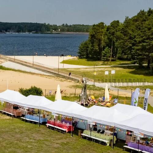 Strefa nad jeziorem z białymi pawilonami targowymi, leżakami przy metalowej rzeźbie, piaszczystą plażą i lasem w tle.