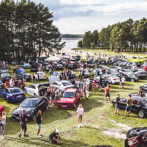 Tłumny zlot samochodowy na trawiastej plaży nad jeziorem, rzędy aut, stoisko z parasolem, ludzie spacerują wśród sosen.