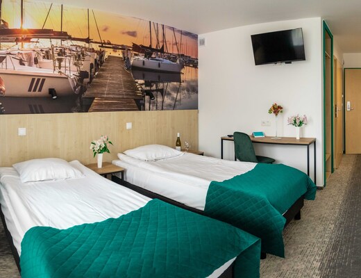 Pokój hotelowy z dwoma pojedynczymi łóżkami, butelką wina na stoliku, biurkiem, telewizorem, fototapetą portu i turkusowymi narzutami.