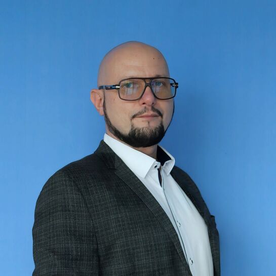 Mężczyzna w eleganckiej marynarce i białej koszuli, z przyciętą brodą i okularami na niebieskim tle.