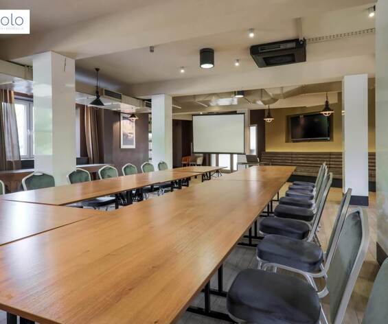 Sala szkoleniowa z U‑kształtnym stołem drewnianym, krzesłami, ekranem projekcyjnym, telewizorem i wiszącymi lampami.