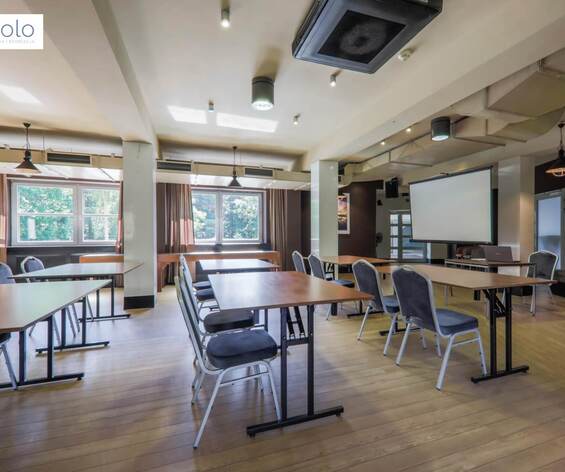 Sala konferencyjna z ustawionymi stołami i krzesłami, ekranem projekcyjnym, dużymi oknami i drewnianą podłogą.