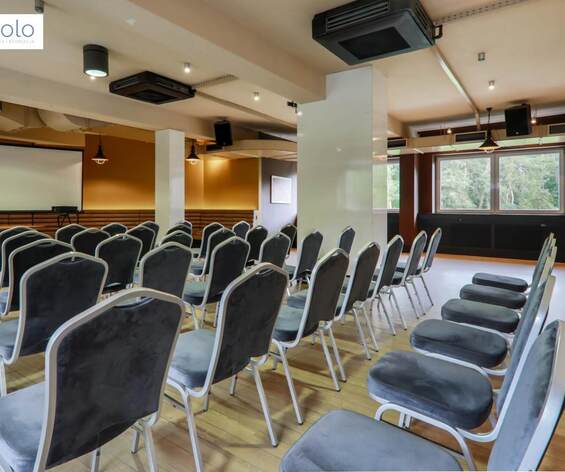 Sala konferencyjna w układzie teatralnym z szarymi krzesłami, ekranem projekcyjnym, nagłośnieniem, drewnianą podłogą i oknami z widokiem na las