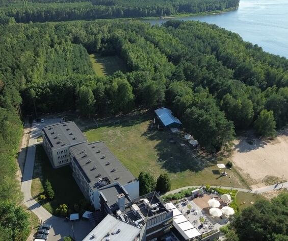 Hotelowy kompleks przy jeziorze: budynek z tarasem i parasolami, piaszczysta plaża, ścieżki i gęsty las