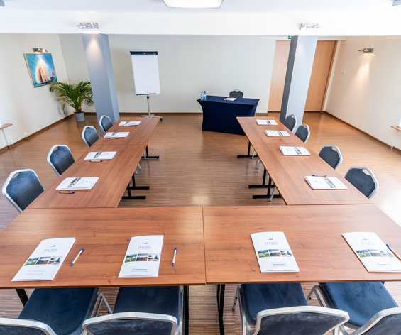 Sala konferencyjna hotelu z układem w U, drewniane stoły, notesy i długopisy, flipchart, stół prelegenta i jasne okna.