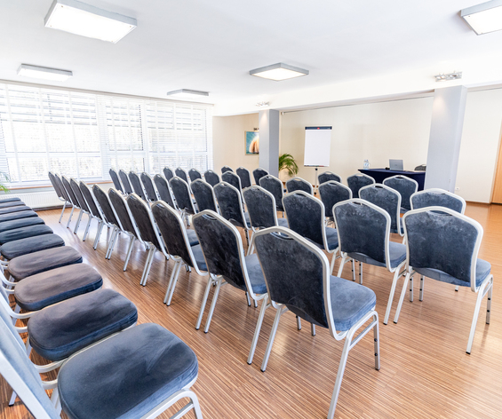 Sala konferencyjna z rzędami szarych krzeseł, dużymi oknami z żaluzjami, flipchartem i stołem prezentera z laptopem i wodą
