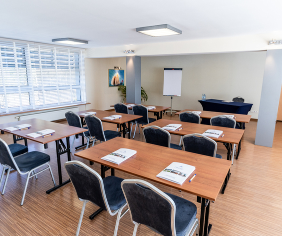 Sala konferencyjna w układzie szkolnym z drewnianymi stolikami, wygodnymi krzesłami, flipchartem i dużymi oknami z żaluzjami.