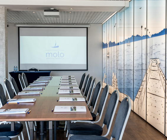 Sala konferencyjna z długim stołem, krzesłami, materiałami, projektorem i świetlną ścianką z ilustracją molo