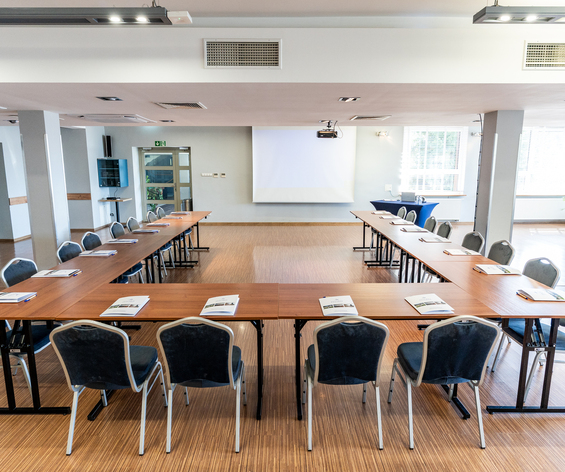 Sala konferencyjna w układzie U z projektorem, drewnianą podłogą, krzesłami i broszurami dla gości
