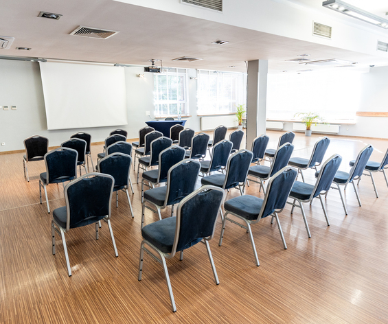 Sala konferencyjna z rzędami niebieskich krzeseł, ekranem projektora, mównicą, dużymi oknami i jasną drewnianą podłogą