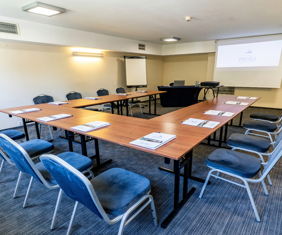 Sala konferencyjna w układzie U z stołami, niebieskimi krzesłami, projektorem, flipchartem i materiałami dla uczestników.
