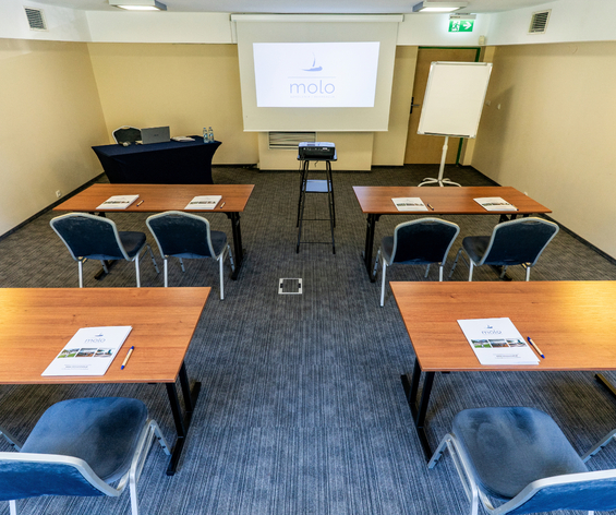 Kameralna sala konferencyjna w hotelu, ustawienie szkoleniowe, projektor i ekran z logo, flipchart, stoliki z notesami.