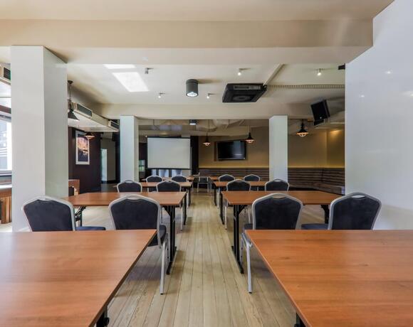 Sala konferencyjna z rzędami stołów i krzeseł, ekranem projekcyjnym, telewizorem, drewnianą podłogą i stonowanym oświetleniem