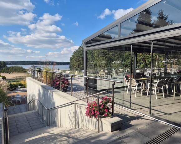 Szklany taras restauracyjny z krzesłami i stolikami, nowoczesna bryła, donice z różowymi kwiatami, widok na plażę i jezioro.