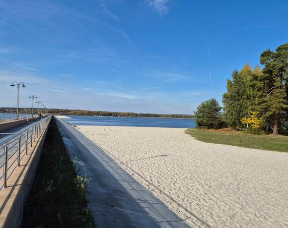 Promenada z lampami i balustradą, goście spacerujący przy piaszczystej plaży nad jeziorem, zalesiony brzeg i spokojna woda