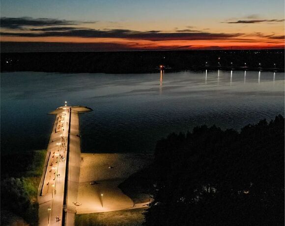 Wieczorny widok z hotelowego balkonu na oświetlony pomost, piaszczystą plażę i spokojne jezioro w barwach zachodu słońca.