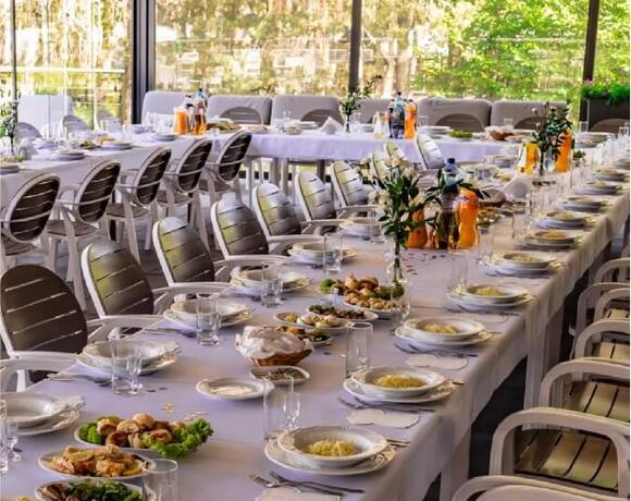 Bankietowy stół w restauracji z białymi obrusami, ustawionymi talerzami, napojami i widokiem na ogród.