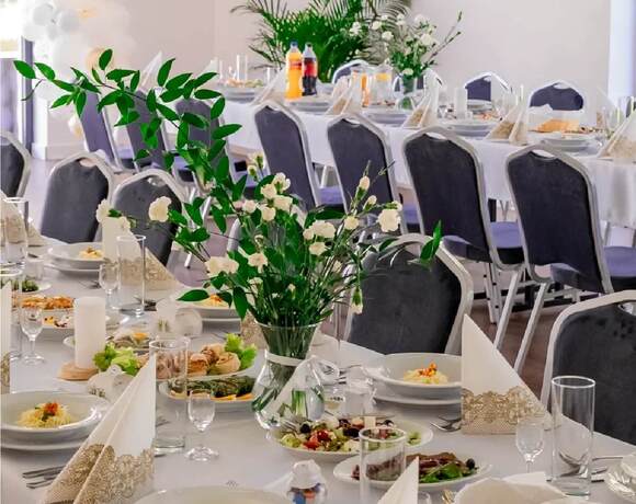 Sala bankietowa na przyjęcie — długie stoły z białymi obrusami, złoto-biała girlanda balonowa, bukiety, eleganckie nakrycia i potrawy.