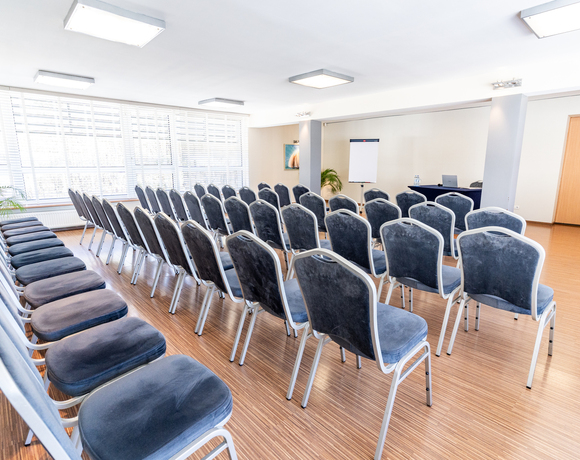 Sala konferencyjna z rzędami szarych krzeseł, dużymi oknami z żaluzjami, flipchartem i stołem prezentera z laptopem i wodą