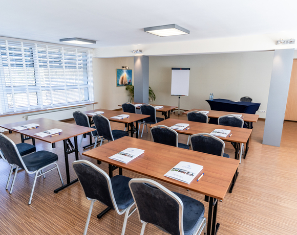 Sala konferencyjna w układzie szkolnym z drewnianymi stolikami, wygodnymi krzesłami, flipchartem i dużymi oknami z żaluzjami.