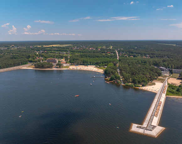 Kurort nad jeziorem z piaszczystą plażą, molem i hotelem przy lesie; kąpieliska, kajaki i leżaki na plaży.