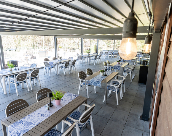 Zadaszony taras restauracyjny ze stolikami, niebieskimi bieżnikami, doniczkami, wiszącymi żarówkami i widokiem na las