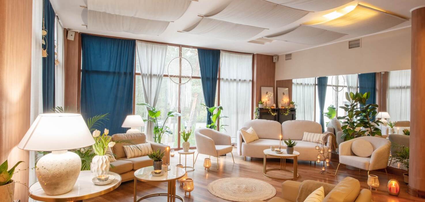 Przytulny hotelowy salon z beżowymi sofami i fotelami, okrągłymi stolikami, roślinami, świecami i dużymi oknami z niebieskimi zasłonami.