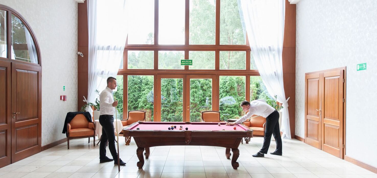 Elegancka sala bilardowa w hotelu: wysokie drewniane okna z widokiem na zieleń, bordowy stół i dwóch grających mężczyzn.