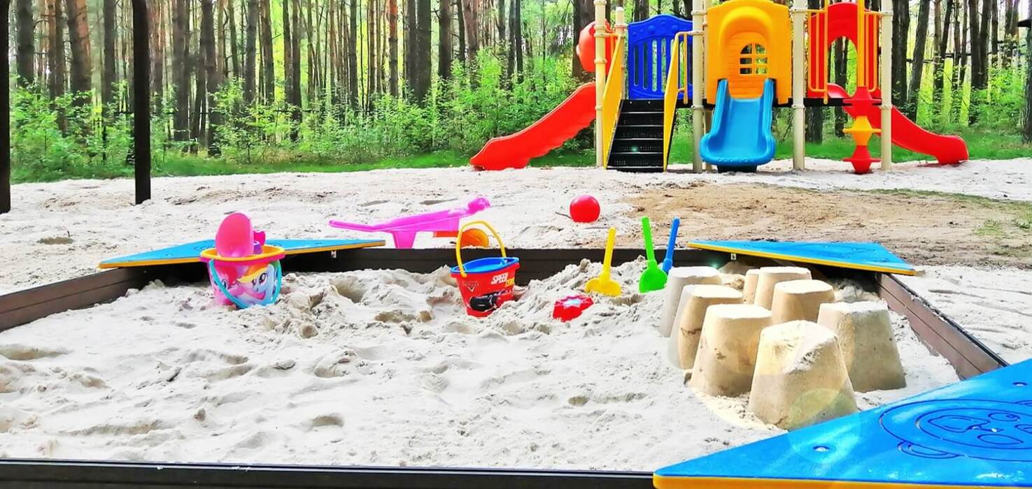 Plac zabaw przy ośrodku: piaskownica z wiaderkami i foremkami oraz kolorowy zestaw zjeżdżalni na skraju sosnowego lasu