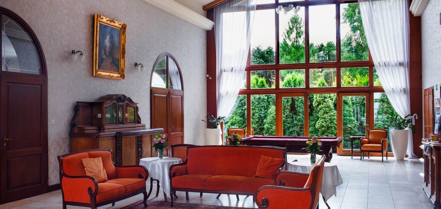 Przestronny hotelowy salon z wysokimi, drewnianymi oknami na ogród, delikatnymi zasłonami, pomarańczowymi sofami, bilardem i klasycznymi meblami.