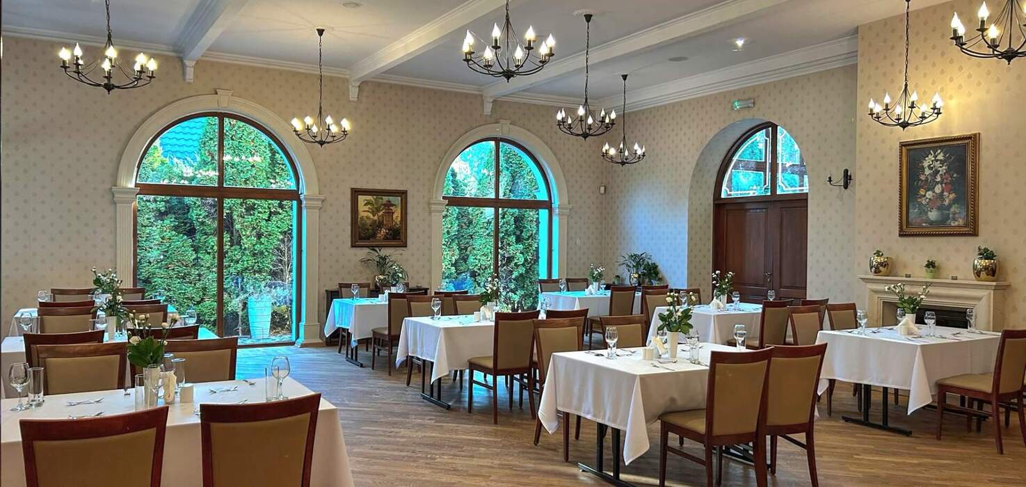 Elegancka jadalnia hotelowa z białymi obrusami, nakrytymi stołami, żyrandolami, dębową podłogą i dużymi półokrągłymi oknami na ogród