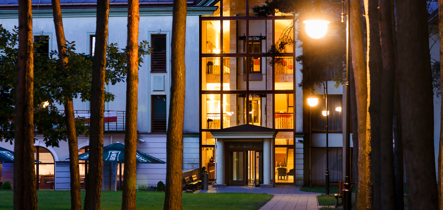 Przeszklone atrium hotelu nocą z ciepłym, oświetlonym lobby; alejka z latarniami i ławkami między sosnami, taras z parasolami