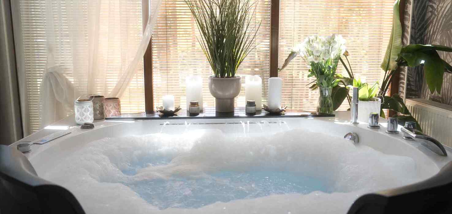 Jacuzzi z bąbelkami w strefie spa, świece, doniczkowe rośliny i naturalne światło przez żaluzje.