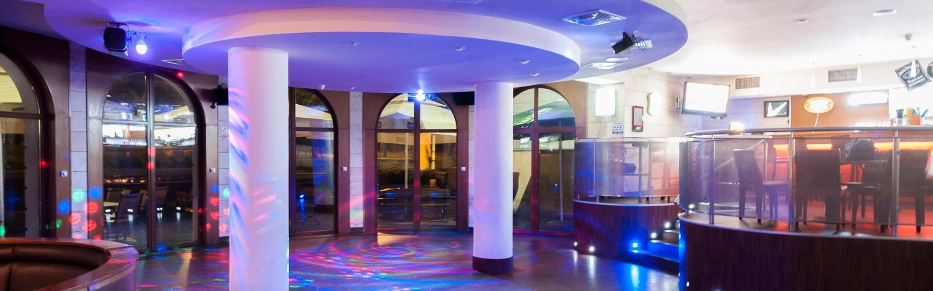 Stylowy bar hotelowy z podwyższonym barem i okrągłym sufitem, filarami oraz parkietem skąpanym w kolorowych światłach