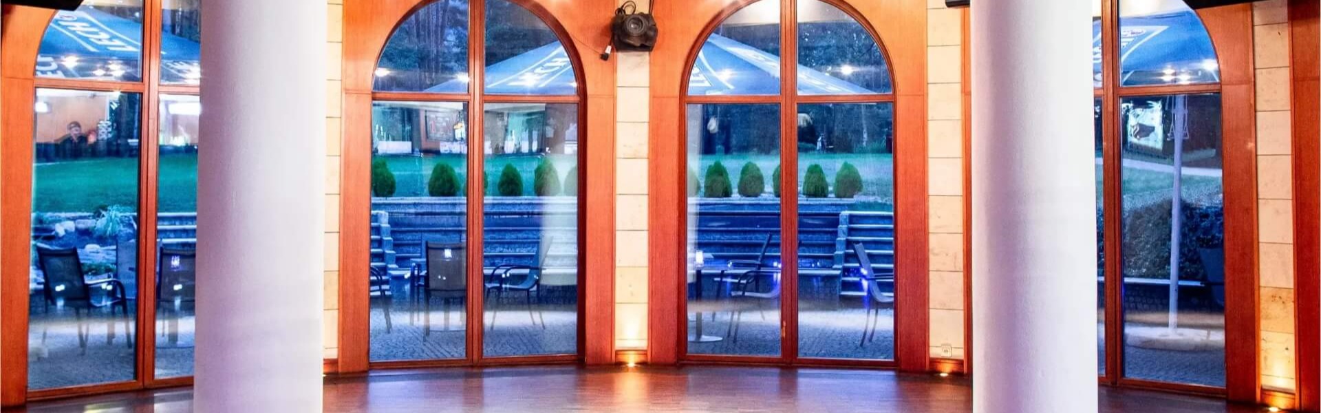 Przestronna sala bankietowa z drewnianą podłogą, kolumnami, sufitem z nagłośnieniem i projektorem, łukowe okna z widokiem na taras