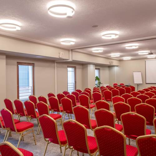 Sala konferencyjna z rzędami czerwonych krzeseł, ekranem i projektorem, flipchartem i jasnym okrągłym oświetleniem
