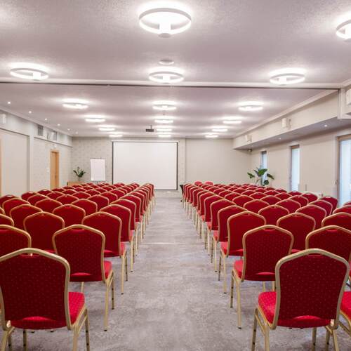 Sala konferencyjna z czerwonymi krzesłami w rzędach, projektorem i ekranem, nowoczesne oświetlenie i neutralne wnętrze