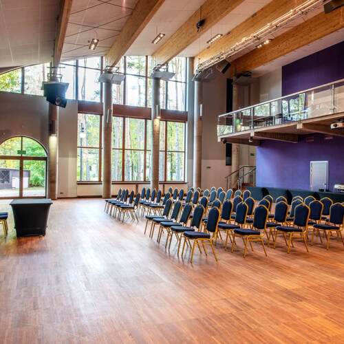 Sala konferencyjna z rzędami krzeseł, wysokimi oknami z widokiem na las, drewnianymi belkami i antresolą.