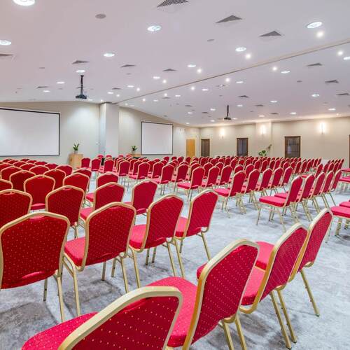 Przestronna sala konferencyjna z rzędami czerwonych krzeseł, dwoma ekranami, projektorem, mównicą i dyskretnym oświetleniem sufitowym