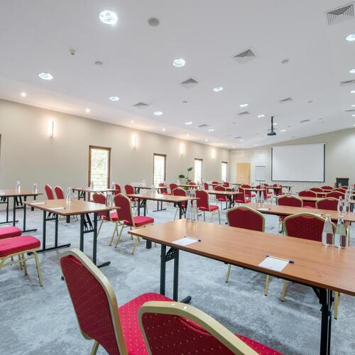 Konferencyjna sala z ustawieniem klasowym, czerwonymi krzesłami, stołami, ekranem projektora, butelkami wody i jasnym oświetleniem