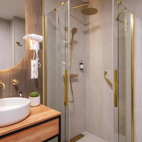 Elegancka łazienka hotelowa z kabiną prysznicową z deszczownicą, złotymi armaturami, umywalką nablatową i suszarką.