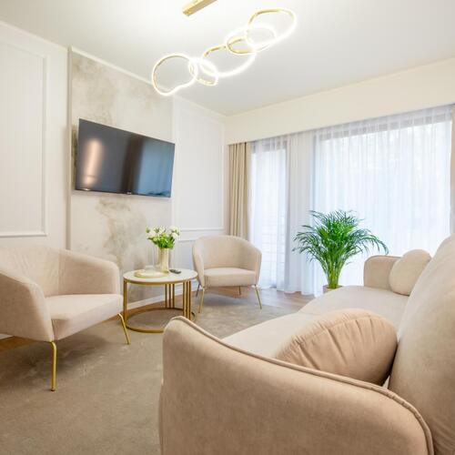 Jasny apartament z beżowymi fotelami i sofą, stolik kawowy z kwiatami, TV na ścianie, nowoczesne oświetlenie