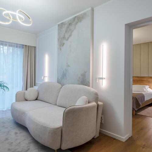 Przestronny salon apartamentu z beżową sofą, designerskim oświetleniem, rośliną i wejściem do przytulnej sypialni